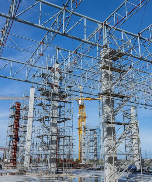 Wide view of scaffolding and steel framework at a muddy construction site, with exposed beams and rebar under clear skies.